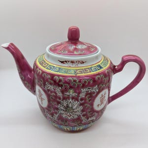 May include: A pink porcelain teapot with a floral design and Chinese characters. The teapot has a lid with a knob on top and a handle on the side.
