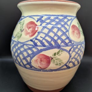 May include: A ceramic vase with a cream body and a blue lattice pattern. The vase features red fruit and green leaf designs. The rim is reddish-brown, and the base is also reddish-brown.