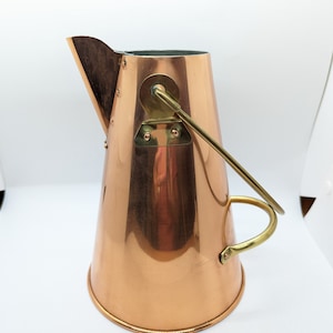 Puede incluir: Jarra de color cobre con asa y detalles en latón. La jarra tiene forma cónica con un pico vertedor y base redondeada. El asa está sujeta al lateral con soportes de latón. La jarra es probablemente vintage y está en buen estado, con unos 20 cm de altura.