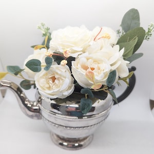 May include: A silver teapot with a bouquet of white roses and greenery inside.