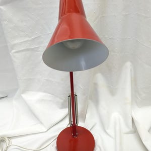K&ouml;nnte beinhalten: Eine rote Schreibtischlampe mit konischem Schirm und rundem Fu&szlig;. Die Lampe hat einen flexiblen Arm mit Federmechanismus und einen wei&szlig;en Innenschirm. Die Lampe ist an einem wei&szlig;en Netzkabel angeschlossen.