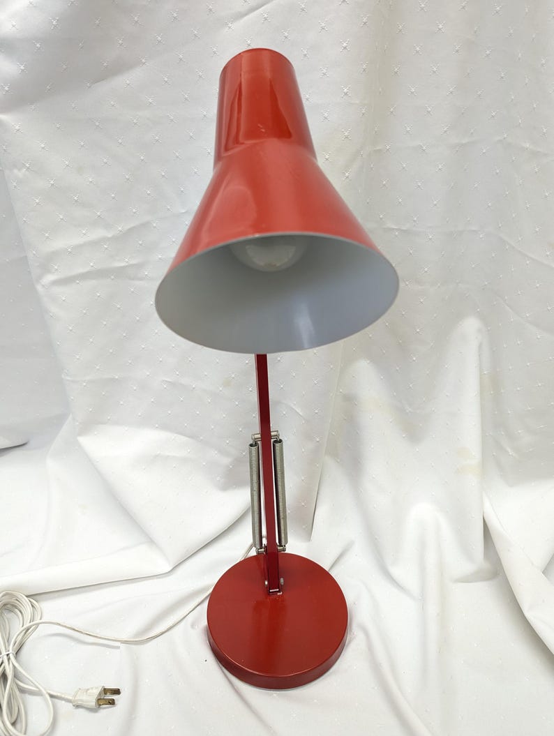 K&ouml;nnte beinhalten: Eine rote Schreibtischlampe mit konischem Schirm und rundem Fu&szlig;. Die Lampe hat einen flexiblen Arm mit Federmechanismus und einen wei&szlig;en Innenschirm. Die Lampe ist an einem wei&szlig;en Netzkabel angeschlossen.
