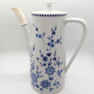 Puede incluir: Cafetera de cerámica blanca con tapa y asa. La cafetera está decorada con motivos florales azules y un pico blanco con punta beige. El borde de la tapa tiene un diseño punteado azul.