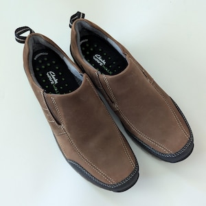 May include: A pair of brown leather slip-on shoes with black soles and white stitching. The shoes have a pull tab on the back and the brand name "Clarks WAVE WALK" is visible on the insole.