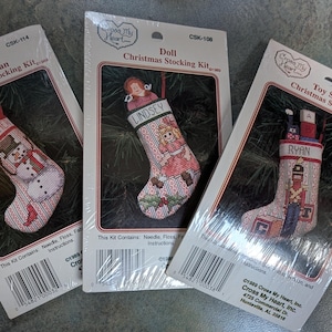 May include: Three Christmas stocking cross-stitch kits. One features a snowman with the word "TEACHER", another a doll with the name "LINDSEY", and the third a toy soldier with the name "RYAN". Each kit includes needle, floss, and instructions.