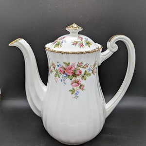 Peut inclure: Théière en porcelaine blanche avec un motif floral composé de roses roses et de feuilles vertes. La théière est ornée d'une bordure dorée autour du couvercle et du bec.