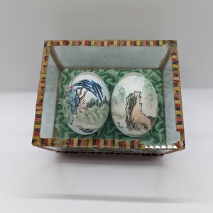 Puede incluir: Dos huevos de porcelana pintados a mano con intrincadas escenas de paisajes, expuestos en una caja con tapa de vidrio, forro de terciopelo verde y borde de rayas de colores.