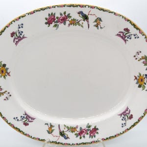 May include: Oval white serving platter with a floral and bird design. The rim features a gold trim and a border of green, pink, and purple flowers. The center of the platter is plain white.