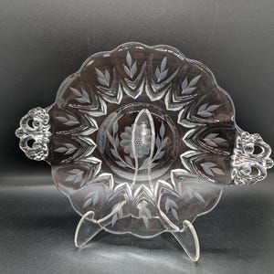 May include: A clear glass serving dish with a scalloped edge and etched leaf and floral designs. The dish has two ornate handles and is supported by a clear stand. The design is symmetrical and elegant.
