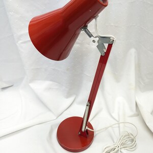 K&ouml;nnte beinhalten: Rote Schreibtischlampe mit konischem Schirm und rundem Fu&szlig;. Die Lampe hat einen schwenkbaren Arm und ein wei&szlig;es Netzkabel. Die Lampe ist aus Metall und hat eine gl&auml;nzende Oberfl&auml;che. Der Sockel hat einen Durchmesser von ca. 15 cm.