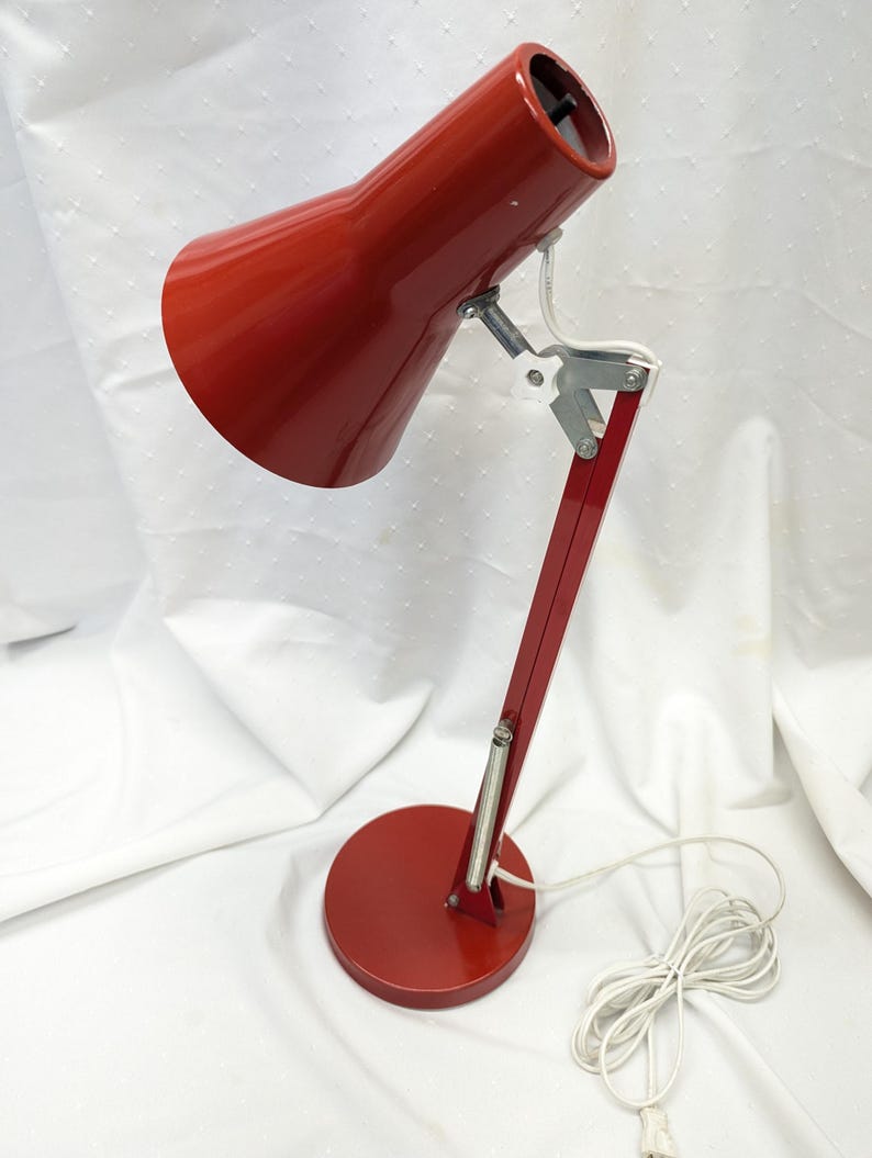 K&ouml;nnte beinhalten: Rote Schreibtischlampe mit konischem Schirm und rundem Fu&szlig;. Die Lampe hat einen schwenkbaren Arm und ein wei&szlig;es Netzkabel. Die Lampe ist aus Metall und hat eine gl&auml;nzende Oberfl&auml;che. Der Sockel hat einen Durchmesser von ca. 15 cm.