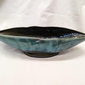 Puede incluir: Un cuenco central de cerámica de color verde azulado y negro. El cuenco tiene forma ovalada y alargada, con un interior oscuro y un exterior verde azulado. El cuenco se apoya sobre una base verde oscuro.