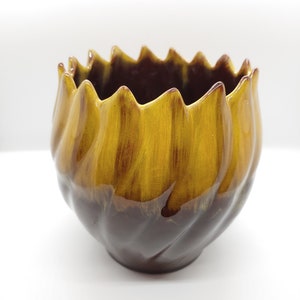 Puede incluir: Un macetero de cerámica marrón y amarillo con un borde ondulado y fruncido. El macetero está esmaltado con un patrón de remolino marrón y amarillo.