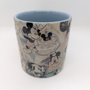 Op de afbeelding: Cilindrische keramische mok met een collage van Mickey Mouse-illustraties in zwart, wit en grijstinten. De binnenrand is lichtblauw. De mok toont Mickey Mouse die een verjaardag viert met een taart.