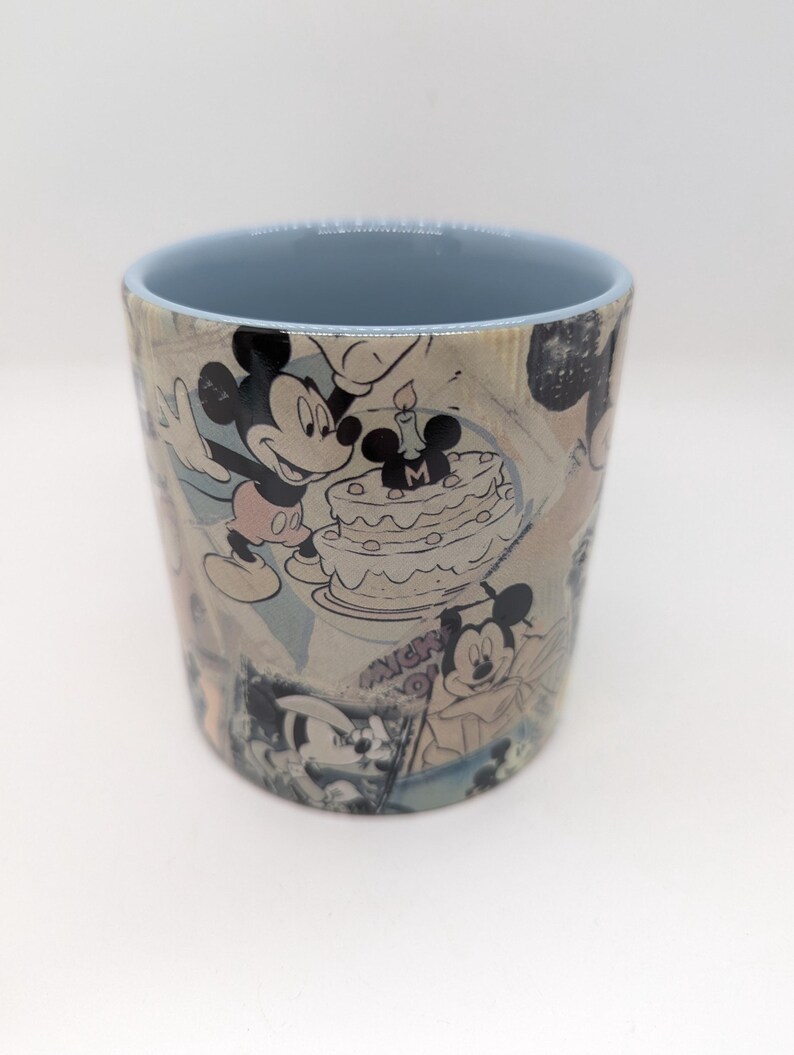 Op de afbeelding: Cilindrische keramische mok met een collage van Mickey Mouse-illustraties in zwart, wit en grijstinten. De binnenrand is lichtblauw. De mok toont Mickey Mouse die een verjaardag viert met een taart.