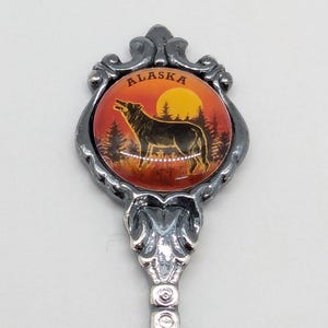 Può includere: Cucchiaio decorativo con un emblema circolare raffigurante la silhouette di un lupo contro un tramonto. L'emblema è incorniciato da un bordo ornato grigio scuro, con la scritta "ALASKA" in alto. Il manico del cucchiaio è argentato.