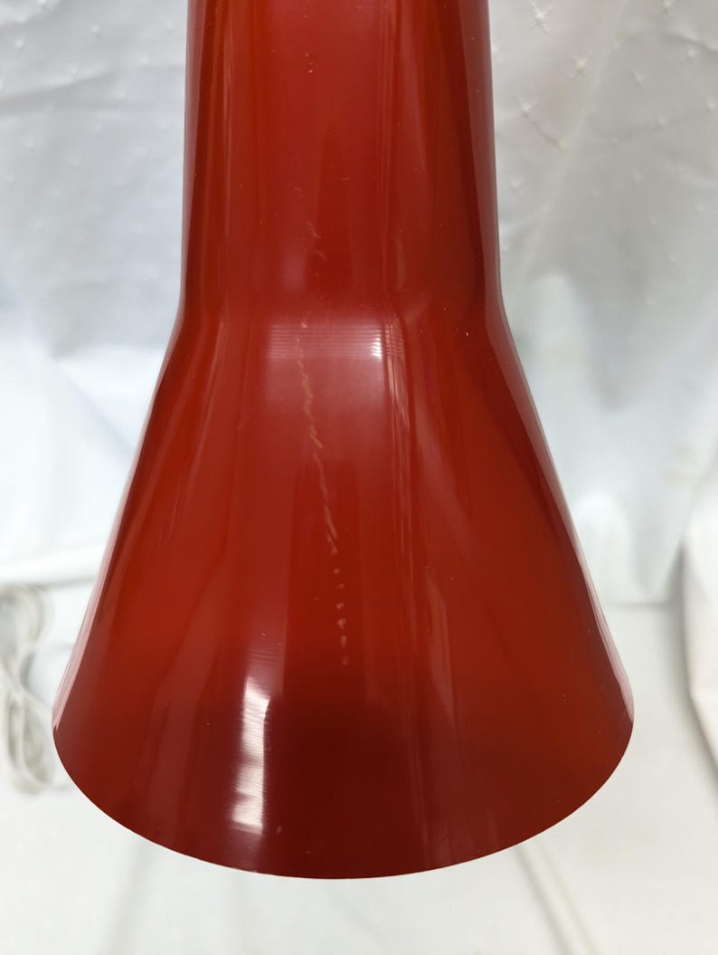 K&ouml;nnte beinhalten: Nahaufnahme eines roten, kegelf&ouml;rmigen Lampenschirms. Der Schirm hat eine gl&auml;nzende Oberfl&auml;che und eine glatte Oberfl&auml;che. Der Lampenschirm hat eine tiefe, satte rote Farbe.