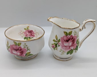 May include: A white porcelain creamer and sugar bowl set. Each piece is decorated with pink roses and green leaves. The rims and handles are trimmed with gold. The sugar bowl has a rounded shape, and the creamer has a handle and spout.