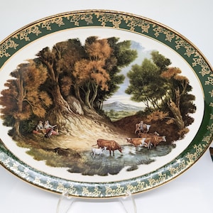 May include: Oval serving platter with a pastoral scene. The center features a landscape with trees, a stream, and cows. The rim is green with a gold floral design and a gold edge.