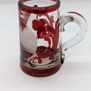 May include: A clear glass mug with a handle, featuring red floral designs and trim. The mug has a cylindrical shape and a clear handle. The red design is visible on the front and sides of the mug. The mug is likely used for drinking.