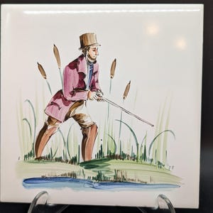 Könnte beinhalten: Quadratische Keramikfliese mit handgemalter Illustration einer Person mit Zylinder und Mantel, die einen langen Stock hält, in der Nähe von Wasser und hohem Gras. Die Fliese hat einen weißen Hintergrund mit glänzendem Finish.