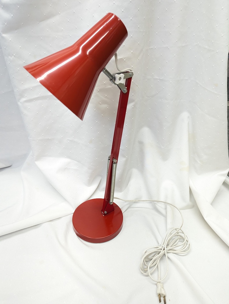 K&ouml;nnte beinhalten: Eine rote Schreibtischlampe mit konischem Schirm und rundem Fu&szlig;. Die Lampe hat einen flexiblen Arm und ein wei&szlig;es Netzkabel. Die Lampe ist aus Metall und hat eine gl&auml;nzende Oberfl&auml;che. Die Lampe ist f&uuml;r den Schreibtischgebrauch konzipiert.
