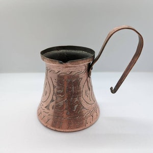Puede incluir: Una pequeña jarra de cobre con base ancha y cuello estrecho, que presenta intrincados patrones grabados a mano en la superficie exterior. El recipiente incluye un asa de metal curvada unida al lateral y un pequeño pico para verter.