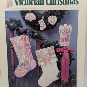 May include: A book cover titled "Victorian Christmas" featuring cross-stitch designs. The image showcases two stockings, an angel figurine, and decorative ornaments, all in soft pinks, whites, and blues. The Dimensions logo is visible at the bottom.