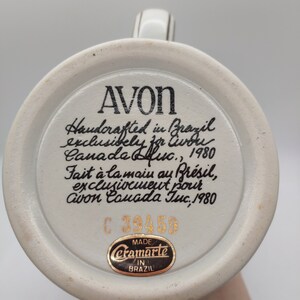May include: A white ceramic mug with a black and gold design. The mug has a handle and a round base. The bottom of the mug has a black and gold sticker that says "Ceramarte Made in Brazil". The mug also has a black and gold sticker with the text "AVON Handcrafted in Brazil exclusively for Avon Canada Inc., 1980 Fait &agrave; la main au Br&eacute;sil, exclusivement pour Avon Canada Inc., 1980 C 39459".