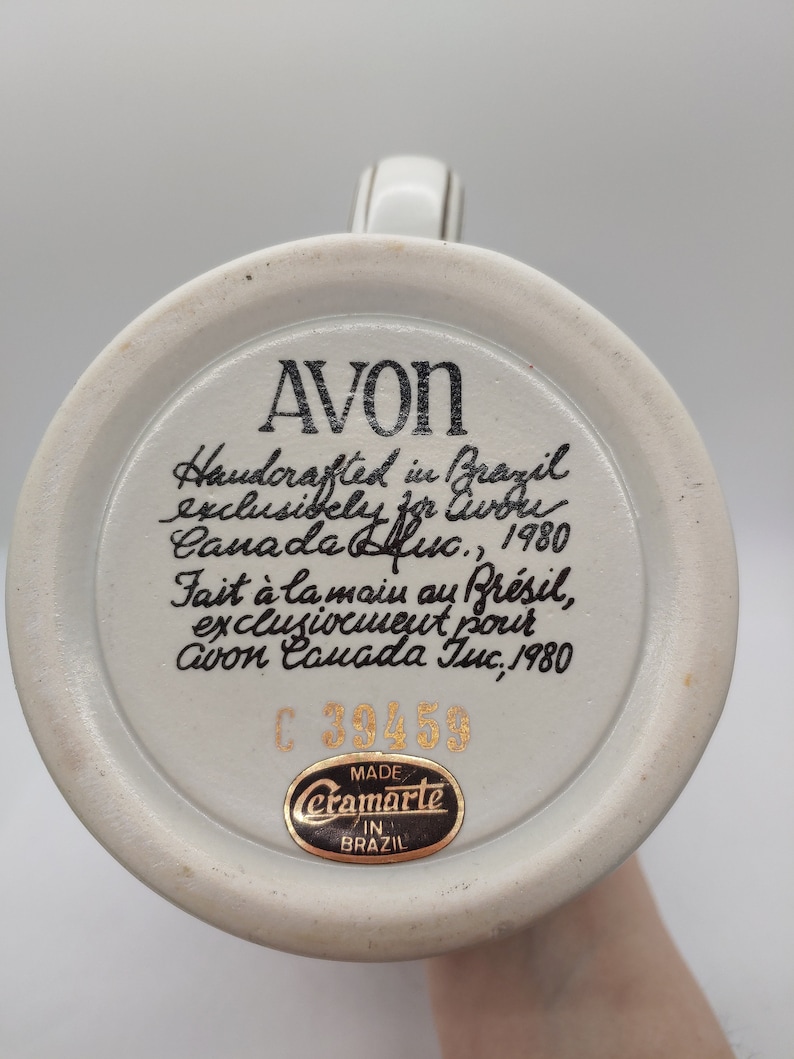 May include: A white ceramic mug with a black and gold design. The mug has a handle and a round base. The bottom of the mug has a black and gold sticker that says "Ceramarte Made in Brazil". The mug also has a black and gold sticker with the text "AVON Handcrafted in Brazil exclusively for Avon Canada Inc., 1980 Fait &agrave; la main au Br&eacute;sil, exclusivement pour Avon Canada Inc., 1980 C 39459".