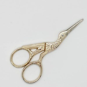 May include: Gold-toned bird-shaped scissors with a detailed design. The scissors have two oval-shaped rings and a pointed tip.