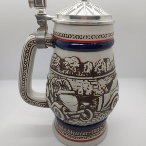 May include: A white ceramic beer stein with a brown and black illustration of a vintage car. The stein has a silver lid and a silver handle with a small silver figure on top. The stein has a blue and red band around the top and the words "Stanley Steamer 1936" on the bottom.