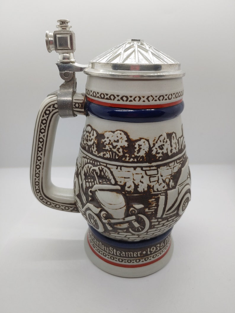 May include: A white ceramic beer stein with a brown and black illustration of a vintage car. The stein has a silver lid and a silver handle with a small silver figure on top. The stein has a blue and red band around the top and the words "Stanley Steamer 1936" on the bottom.
