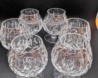 Puede incluir: Seis copas de brandy de cristal transparente con patrones tallados en diamante. Las copas tienen forma de cuenco redondeado y se asientan sobre un tallo corto y una base circular. Las copas están dispuestas en círculo.