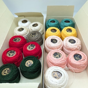 May include: Two open cardboard boxes filled with colorful DMC pearl cotton thread balls. The colors include white, cream, red, green, pink, yellow, and teal. Each ball has a label in the center. The boxes are on a white surface.