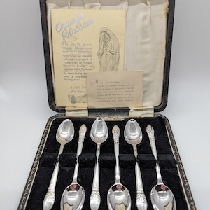 May include: A set of six silver teaspoons in a black velvet-lined box. The spoons are in a traditional design with a floral pattern. The box has a label that reads "Orange Blossom" and includes a description of the product.