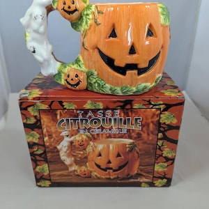 Peut inclure: Une tasse en céramique en forme de citrouille d'Halloween avec une anse représentant un fantôme blanc et une petite citrouille. La tasse repose sur une boîte décorée de feuilles d'automne avec le texte TASSE CITROUILLE EN CÉRAMIQUE.