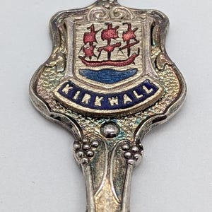 May include: Close-up of a silver souvenir spoon with a decorative handle. The spoon features a shield-shaped emblem with a ship and the word "KIRKWALL" in blue lettering. The emblem has red, blue, and silver accents.