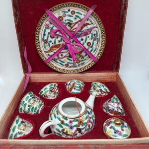 Puede incluir: Un juego de té de porcelana en miniatura en una caja roja con forro de terciopelo rojo. El juego incluye una tetera, seis tazas, seis platillos y un azucarero con tapa. La porcelana está decorada con un patrón floral e imágenes de mujeres con ropa tradicional.