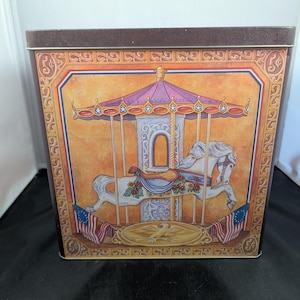 May include: A square metal tin featuring a carousel horse illustration on a golden background. The design includes a purple canopy, a white horse with a blue saddle, and American flag accents. The border displays the text 5 cents in the corners.