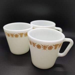 Può includere: Tre tazze in ceramica bianca con disegni floreali marroni intorno al bordo superiore. Le tazze hanno una forma arrotondata e un manico.