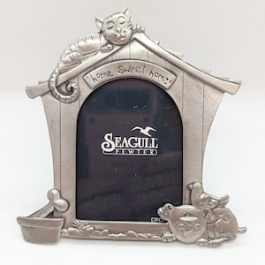 Puede incluir: Un marco de fotos de peltre plateado con forma de caseta de perro con un gato durmiendo en el techo. El marco dice "home sweet home" y tiene un perro y un pájaro en la parte inferior. El marco está hecho por Seagull Pewter.