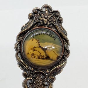 May include: Vintage souvenir spoon with an ornate handle and a circular emblem featuring a polar bear and cubs. The emblem is set against a landscape with the word "ALASKA" above. The spoon has a detailed, antique design.
