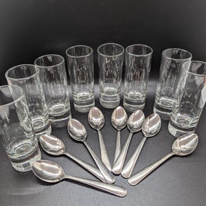 Può includere: Un set di dieci bicchieri da shot in vetro trasparente e dieci cucchiaini d'argento disposti in cerchio su una superficie nera.