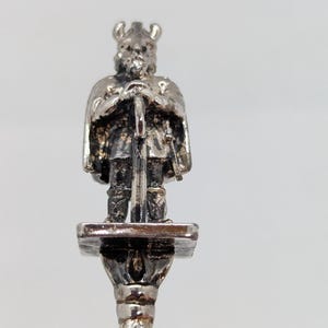 May include: A silver-toned spoon featuring a detailed miniature Viking figure holding a sword. The Viking is standing on a small platform above a fluted column, creating a decorative and historical design. The spoon's handle has a vertical, ribbed pattern.