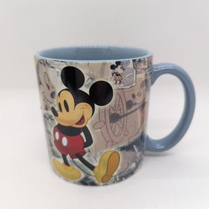 Op de afbeelding: Een keramische mok met Mickey Mouse. De mok heeft een lichtblauw handvat en rand. Mickey Mouse is afgebeeld in zijn klassieke outfit, met een rode broek, gele schoenen en witte handschoenen. De achtergrond heeft een vintage design.