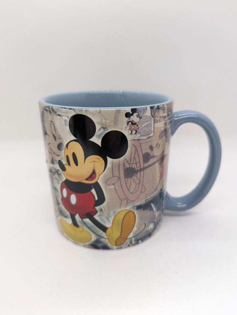 Op de afbeelding: Een keramische mok met Mickey Mouse. De mok heeft een lichtblauw handvat en rand. Mickey Mouse is afgebeeld in zijn klassieke outfit, met een rode broek, gele schoenen en witte handschoenen. De achtergrond heeft een vintage design.