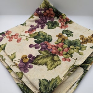 May include: Two folded beige fabric napkins with a grapevine pattern. The pattern features green leaves, purple, red, and green grapes.