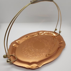 Puede incluir: Una bandeja de servir de cobre con un diseño adornado y un asa de alambre dorado. La bandeja tiene un borde festoneado y un asa de resorte.