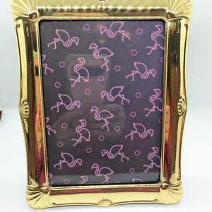 May include: A gold-colored picture frame with a decorative, scalloped edge. The frame has a black background with a pink flamingo pattern.